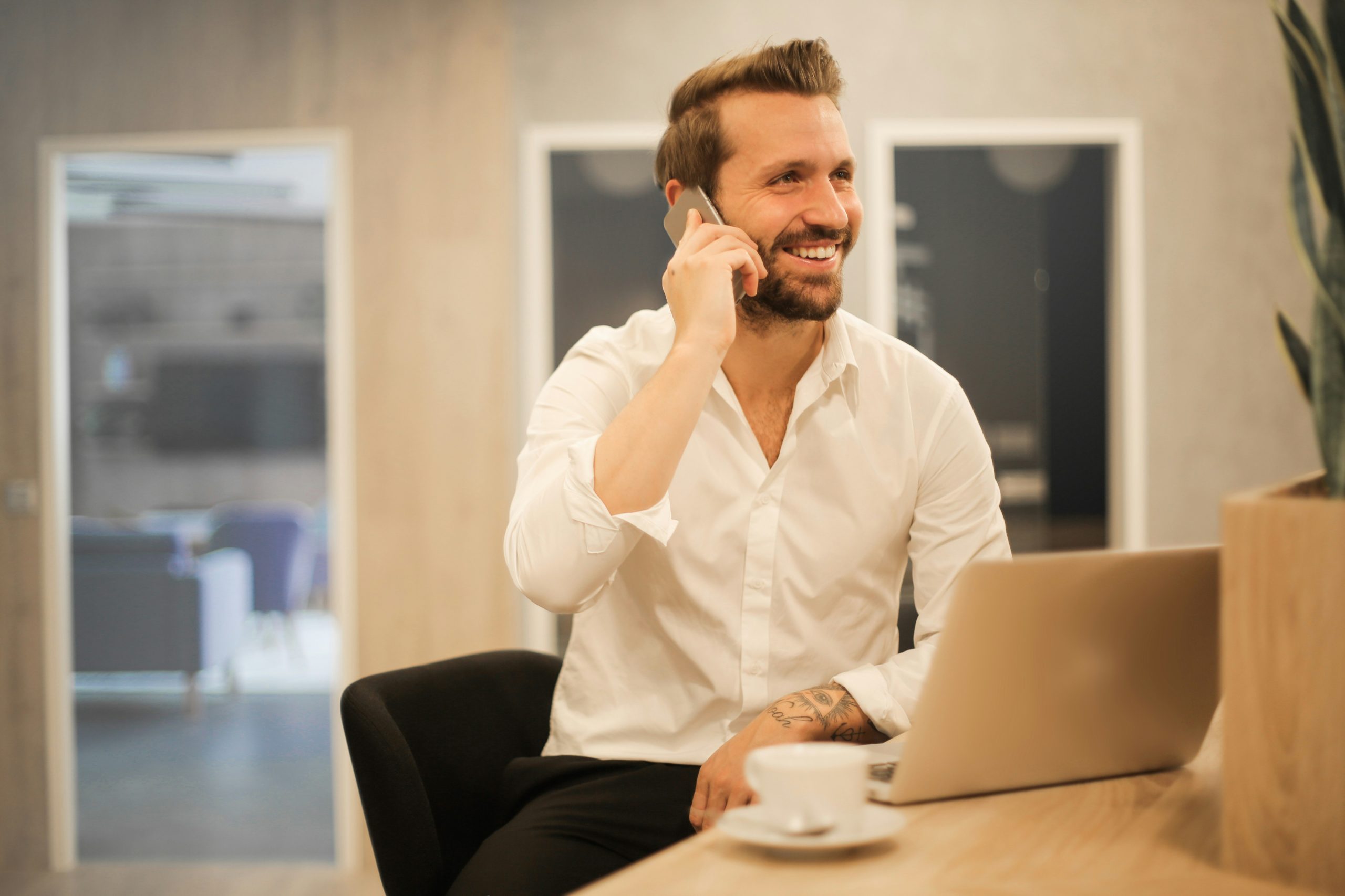 A smiling man dressed in business casual attire working on a laptop while talking on the phone in a cafe and office space.