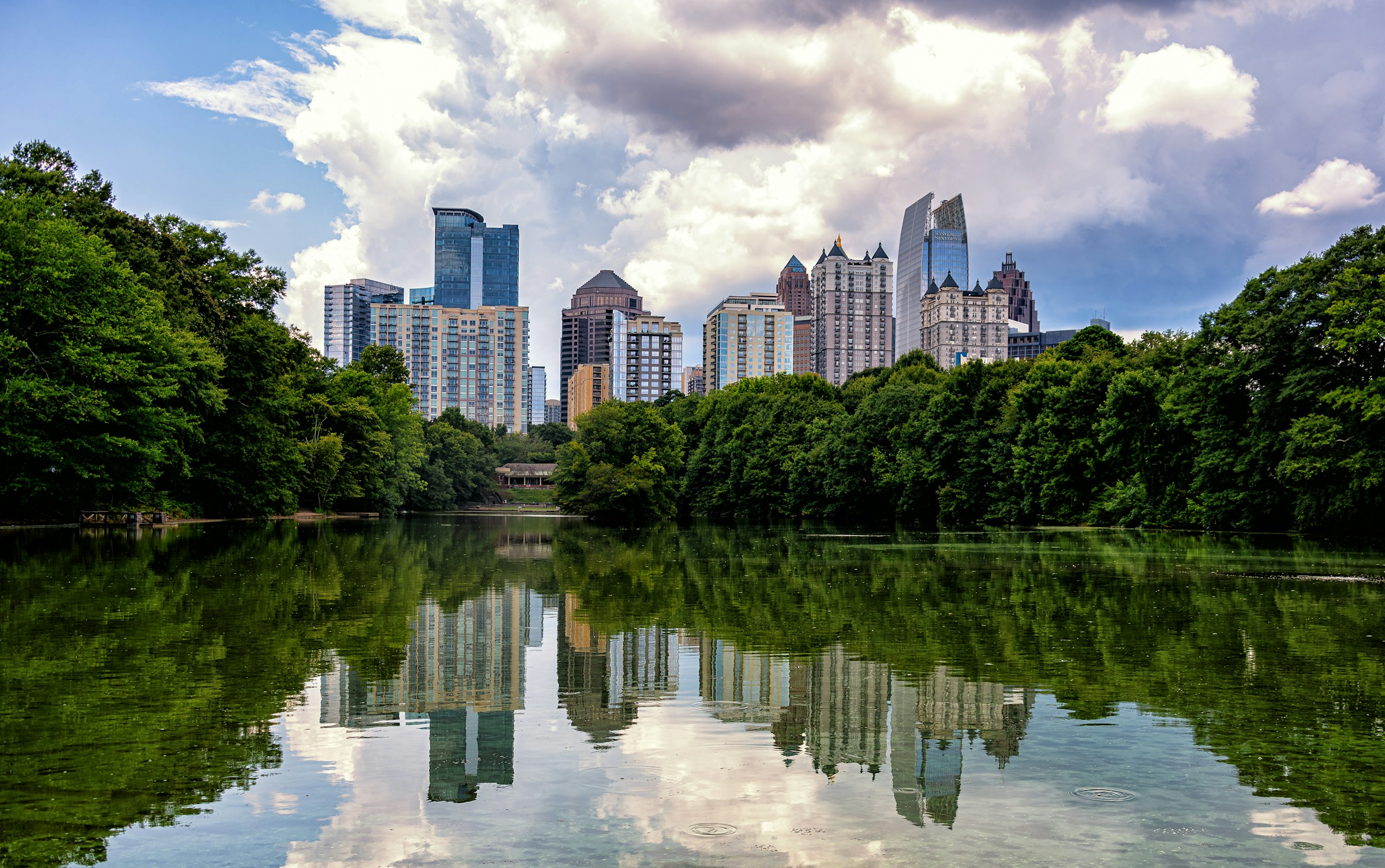 A view of the Atlanta skyline from Piedmont park on a sunny summer day, with the skyline reflected in the park's tree-lined pond.