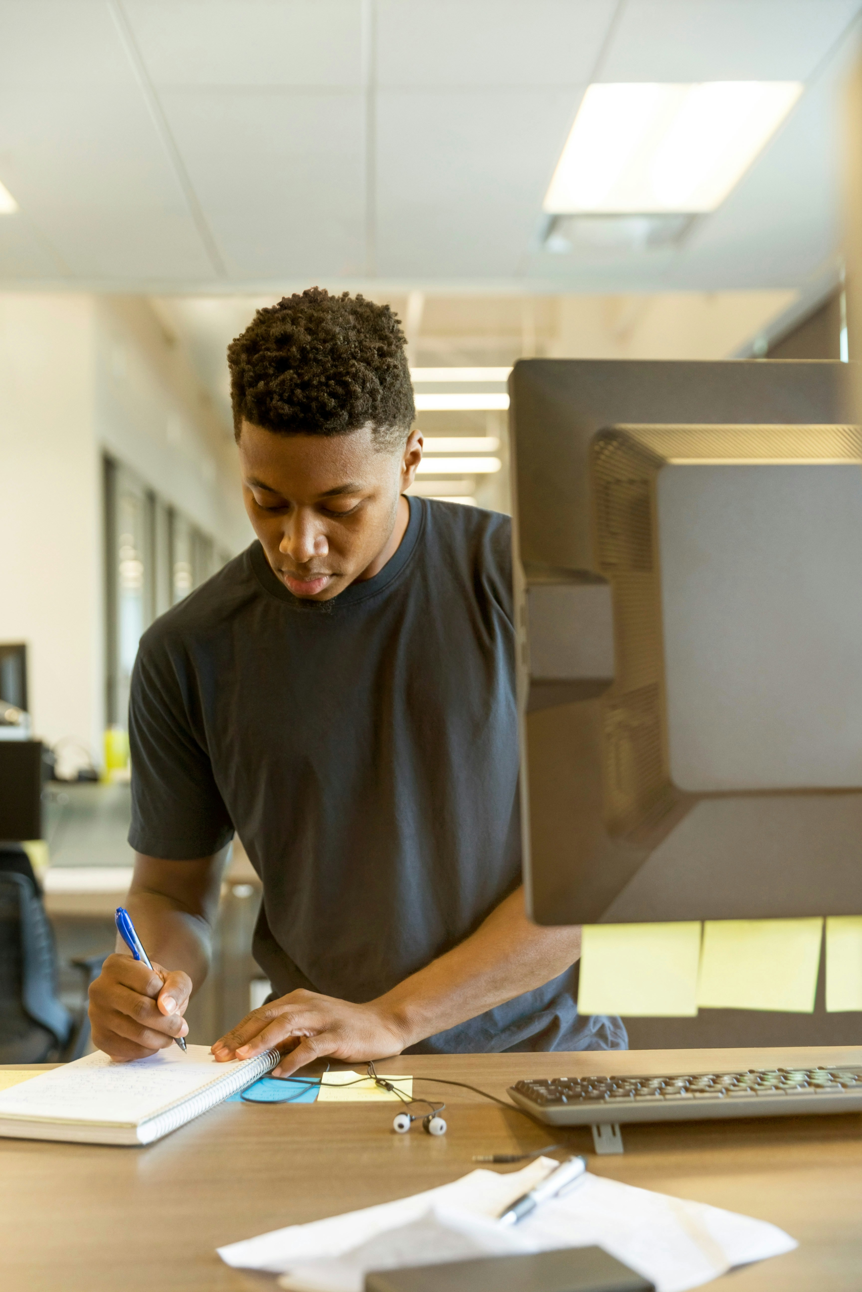 A man writing working at a standing desk, writing in a notebook with a computer in front of him, along with other notes and some earbuds.