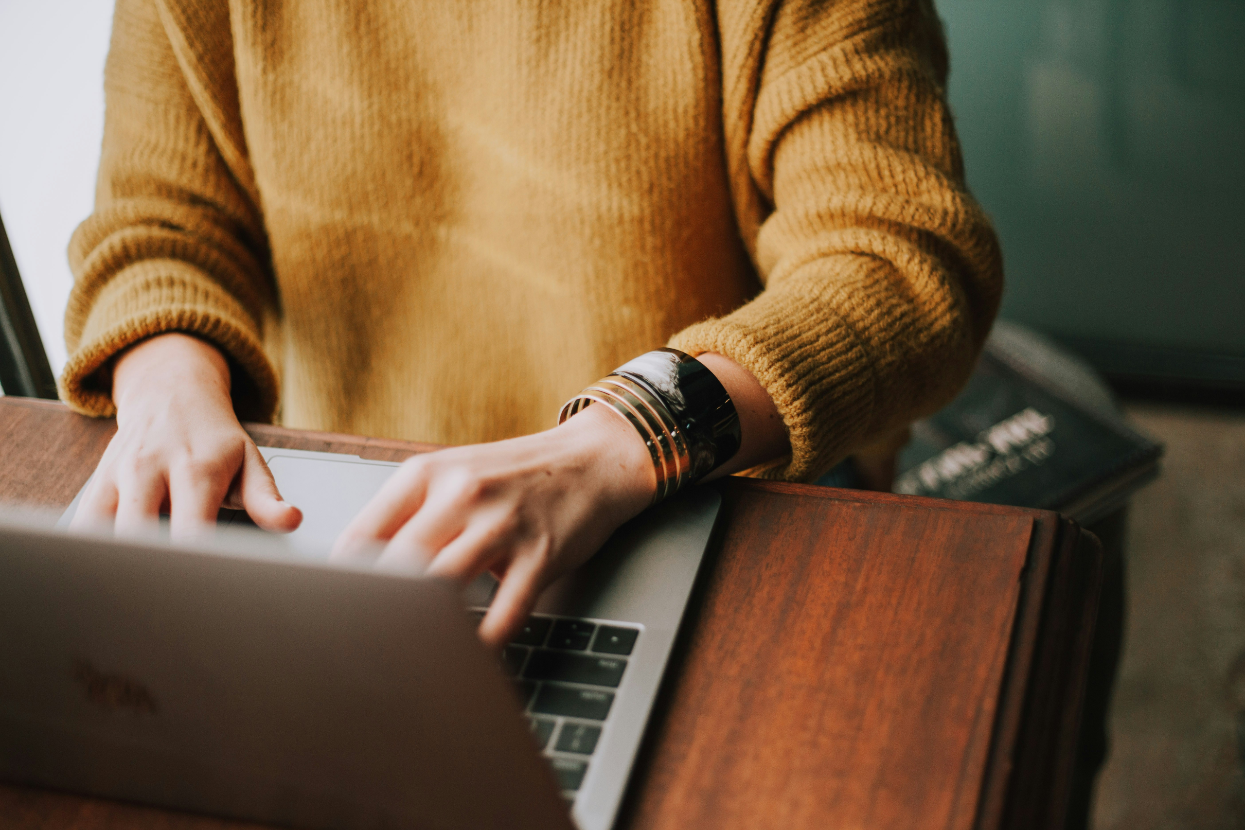 A woman in an orange sweater typing on a laptop, picture from the neck down with the focus on her arms.