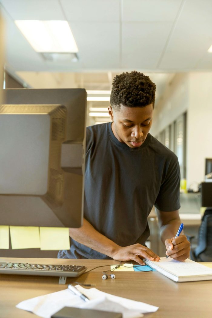 A man writing working at a standing desk, writing in a notebook with a computer in front of him, along with other notes and some earbuds.